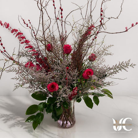 Tall clear glass vase arrangement with red roses, twisting bare branches, red berry-like accents, pale airy filler flowers, and glossy green leaves displayed on a sunlit tiled floor.