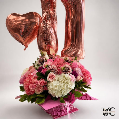 Lush pink and white floral arrangement with mauve flowers, greenery, patterned wrapping, ribbons, and rose gold foil balloons.