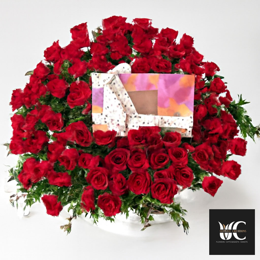 Large round arrangement of red roses with greenery and a decorative rectangular card in the center.