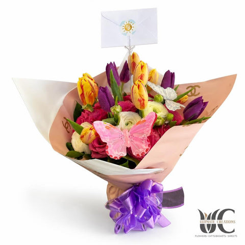 Colorful hand-tied bouquet with pink, purple, yellow, and white flowers, wrapped in peach and white paper with a purple ribbon and butterfly accent.