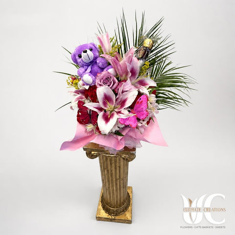 Tall pink and white floral arrangement with a purple teddy bear, lavender rose, red flowers, greenery, pink wrapping, and an ornate gold-toned pedestal vase.
