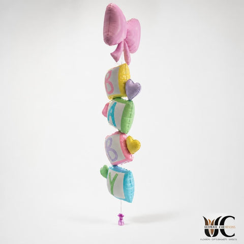 Vertical cluster of pastel foil balloons in pink, green, blue, lavender, and yellow inside a retail store window area, with card racks and candy displays nearby.