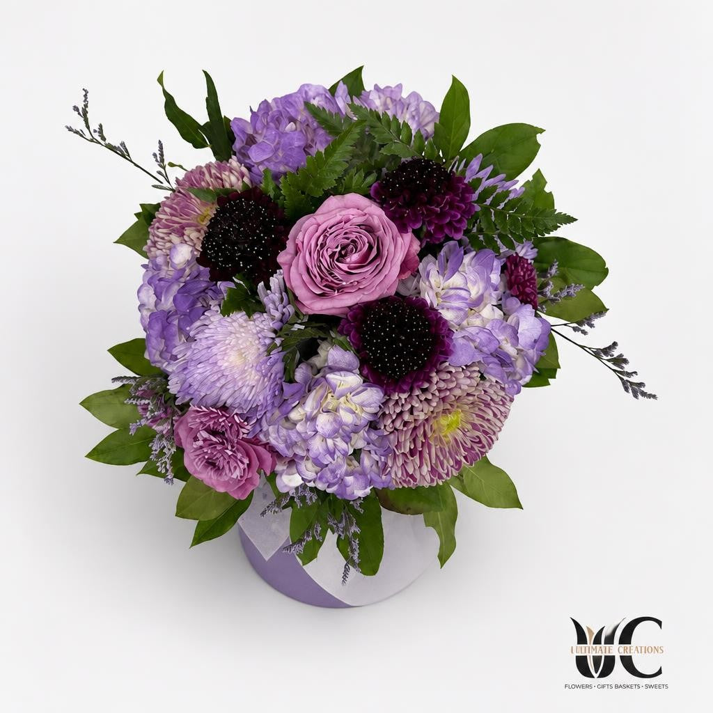 Rounded purple and mauve floral arrangement with dark-centered blooms, textured flowers, and lush green foliage photographed from above.
