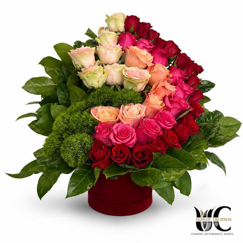 A red flower box filled with layered roses in red, pink, blush, and peach tones with lush green foliage.