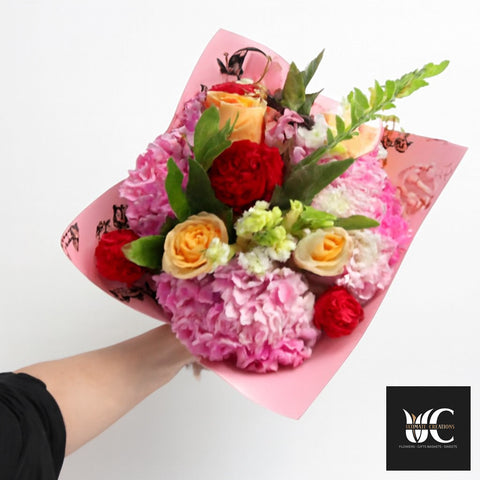 Hand-tied bouquet with pink flowers, peach roses, red blooms, and green foliage wrapped in pink paper.