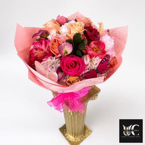 Lush multicolored flower bouquet with pink, blush, coral, peach, and burgundy blooms, wrapped in pink printed paper with a bright pink ribbon and decorative butterfly accent, displayed in a tall gold pedestal vase.