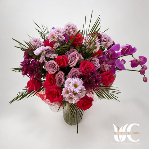 Lush mixed bouquet with red and lavender roses, pink daisy-like chrysanthemums, dark burgundy blooms, purple orchids, and long green tropical leaves displayed indoors.
