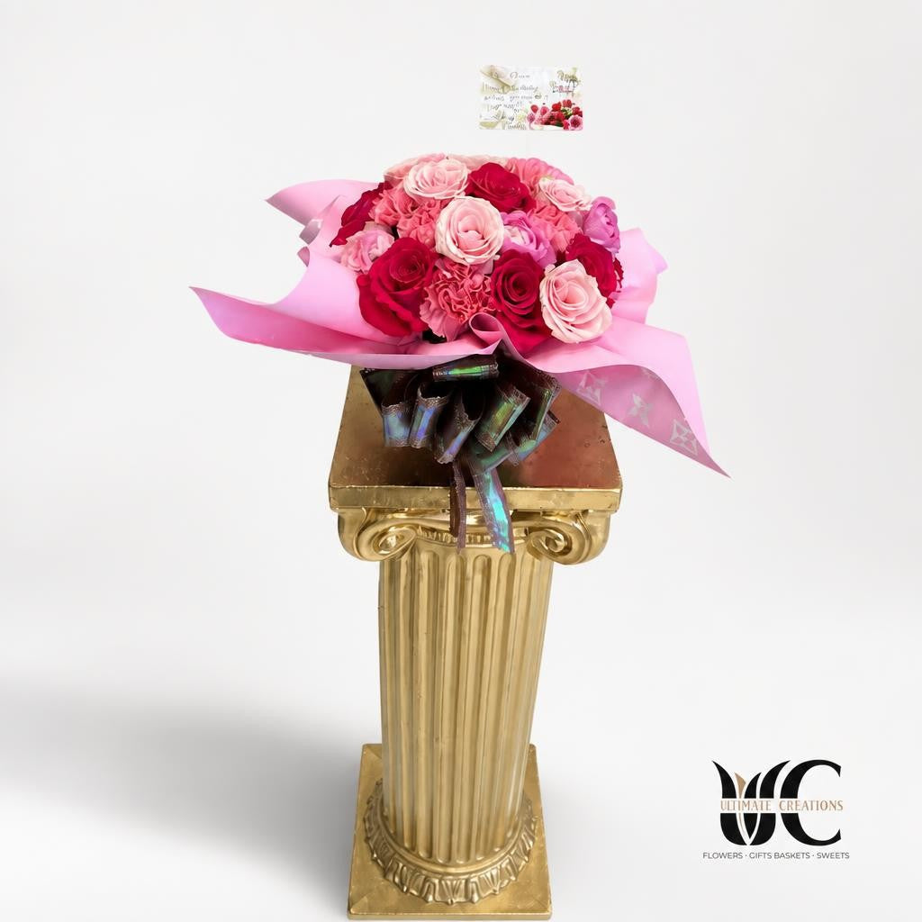 Rounded bouquet of pink and deep red flowers wrapped in bright pink paper and tied with a ribbon, displayed on an ornate gold pedestal in front of a balloon display.