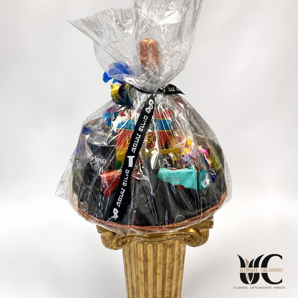Large cellophane-wrapped gift basket filled with assorted packaged treats and colorful items, tied with a black ribbon bearing Hebrew lettering, displayed on an ornate gold pedestal.