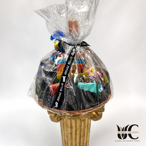Large cellophane-wrapped gift basket filled with assorted packaged treats and colorful items, tied with a black ribbon bearing Hebrew lettering, displayed on an ornate gold pedestal.