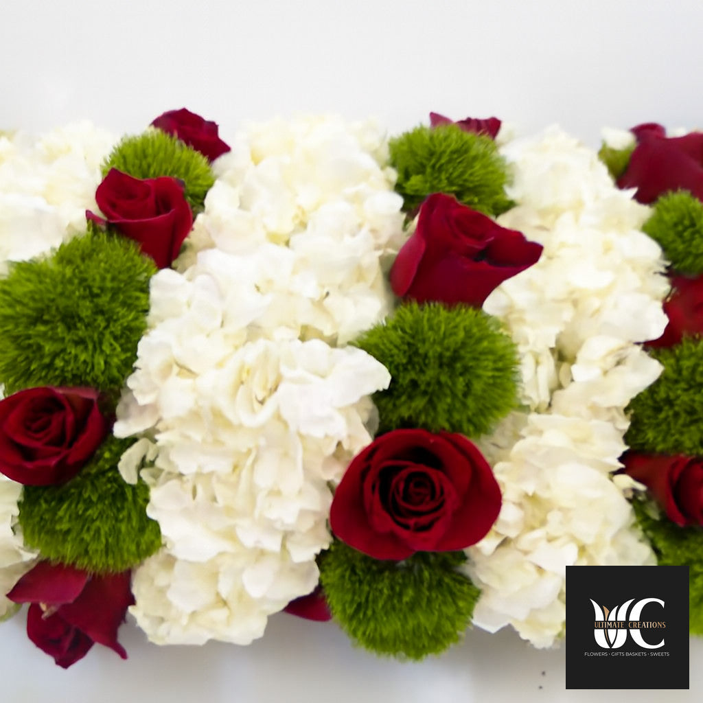 Close-up of a floral centerpiece with red roses, white flowers, and green accent blooms arranged in a dense rectangular design.