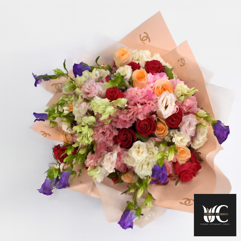Colorful hand-tied bouquet with red, pink, peach, cream, green, and purple flowers wrapped in blush paper.