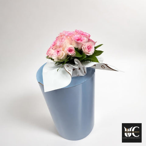 Rounded arrangement of pale pink roses with green leaves in a tall light-blue vessel and white wrapping.
