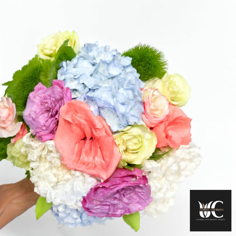 Handheld bouquet of mixed pastel flowers including blue, pink, coral, cream, and green blooms.