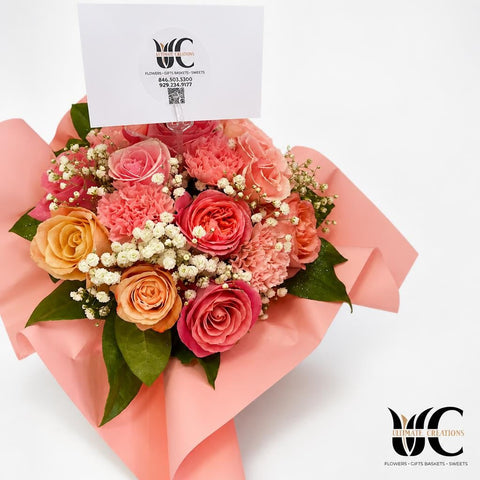 Hand-tied bouquet of pink, peach, coral, and cream flowers with white filler blossoms and green leaves, wrapped in pale pink paper and displayed with a card.