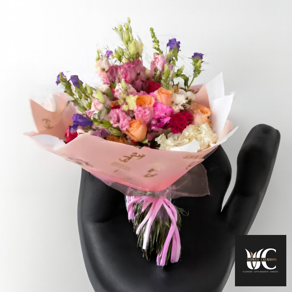 Hand-tied bouquet of pink, peach, purple, and white flowers wrapped in blush paper and ribbon.