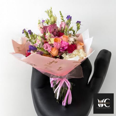 Hand-tied bouquet of pink, peach, purple, and white flowers wrapped in blush paper and ribbon.