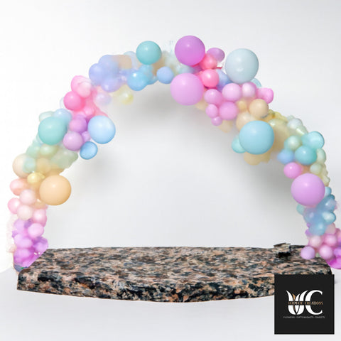 Pastel balloon arch in pink, lavender, blue, mint, and peach over a stone base.