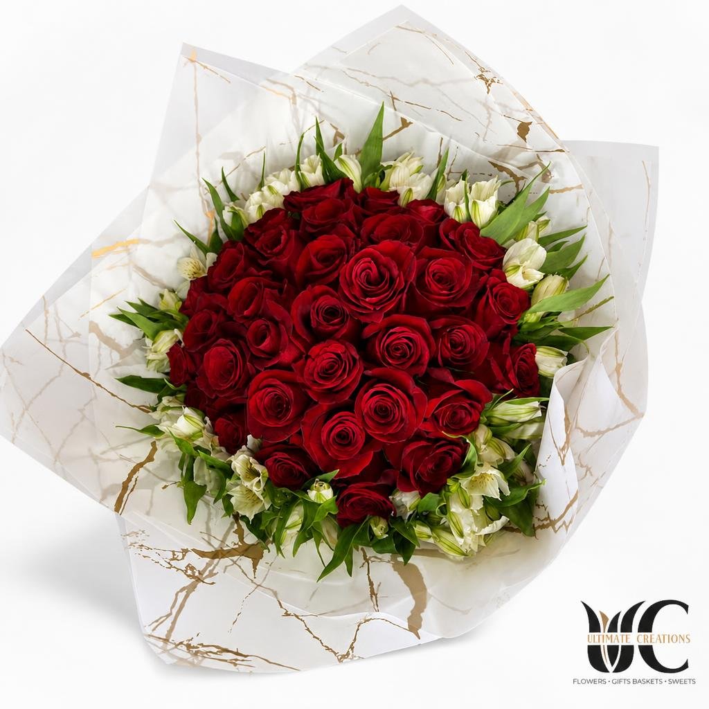 A large bouquet of red roses with white accent flowers and greenery, wrapped in white paper with gold veining.
