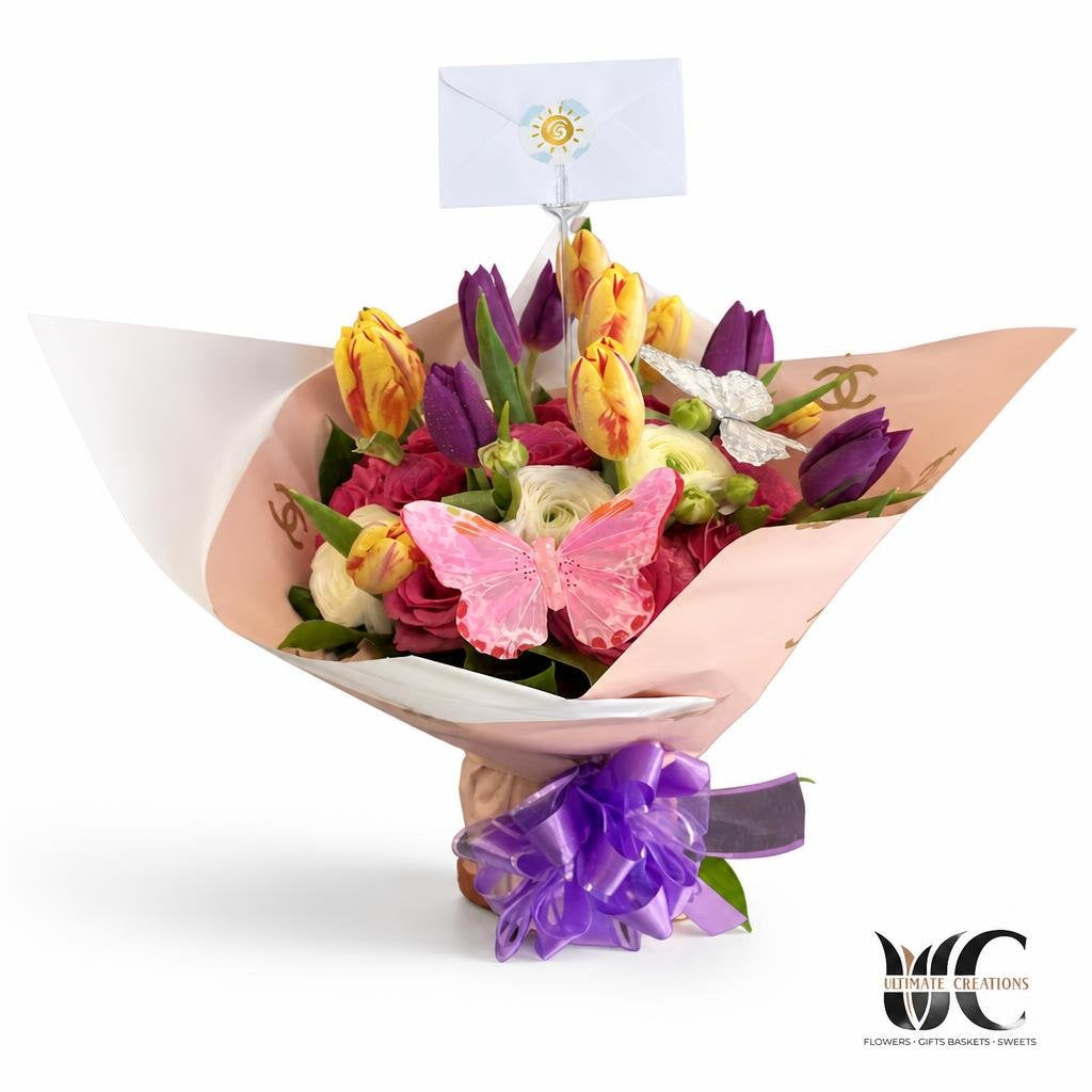 Hand-tied bouquet with pink, purple, yellow, and white flowers wrapped in blush paper with a purple ribbon and butterfly decoration.