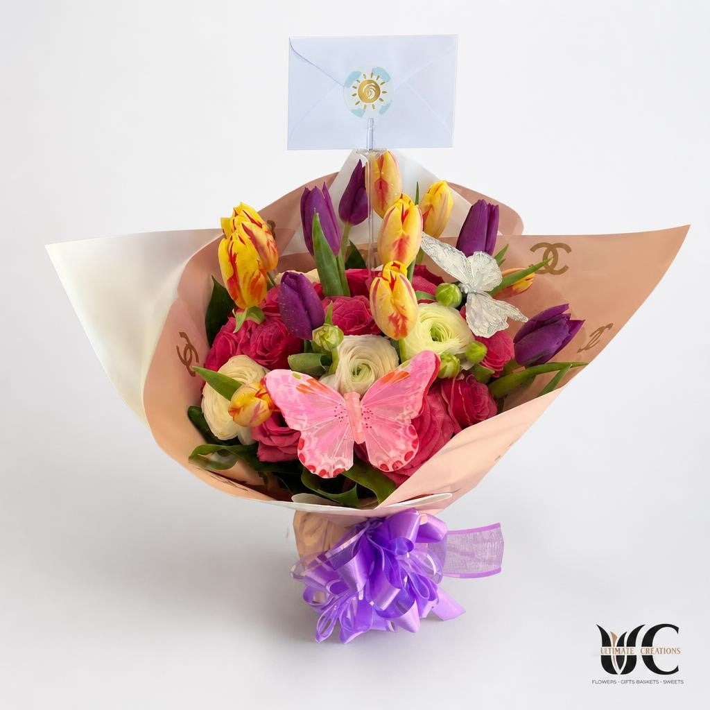 Colorful hand-tied flower bouquet in blush pink wrapping with a purple bow, pink and purple blooms, yellow-and-pink flowers, cream flowers, green foliage, butterfly decorations, and a small envelope card.