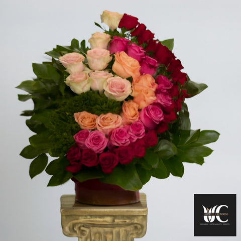 A spiral arrangement of roses in red, pink, peach, and blush with lush green leaves in a vase.