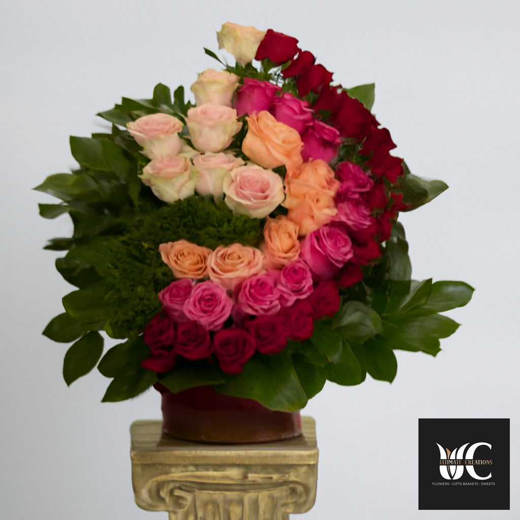 A spiral arrangement of roses in red, pink, peach, and blush with lush green leaves in a vase.