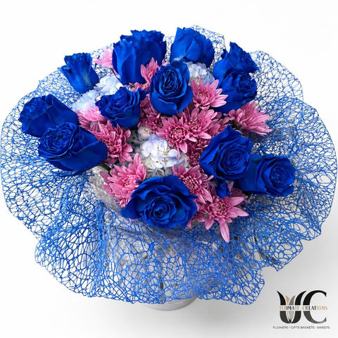 Bouquet of vivid blue roses, pink daisy-like flowers, pale blue-white clustered blooms, and blue netted wrapping.