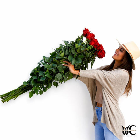Woman holding an oversized bouquet of long-stem red roses with lush green leaves against a light background.