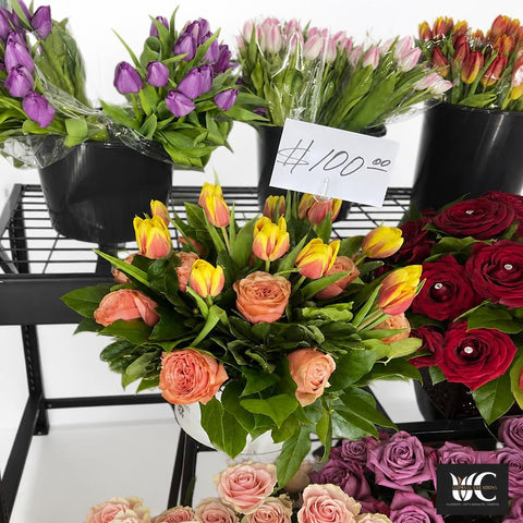 Lush bouquet with peach roses, yellow and coral tulips, green foliage, and surrounding purple and red roses on a flower shop display.