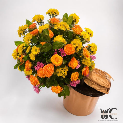 Full rounded floral arrangement with yellow daisy-like flowers, orange blooms, magenta-purple accents, and glossy green leaves in a tall neutral-toned vase on a counter.