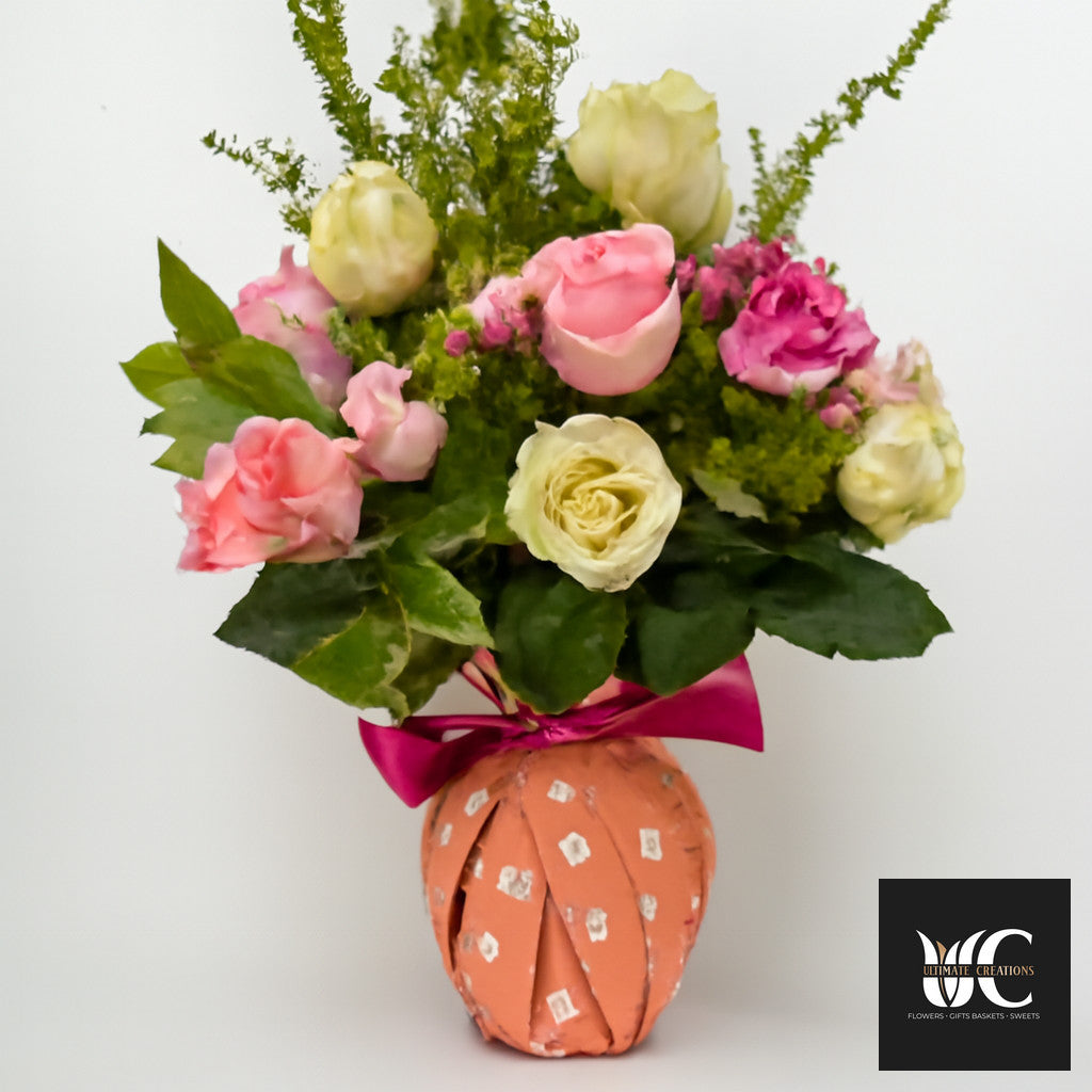 Pink and cream rose arrangement with greenery in a decorative wrapped vase tied with a magenta ribbon.