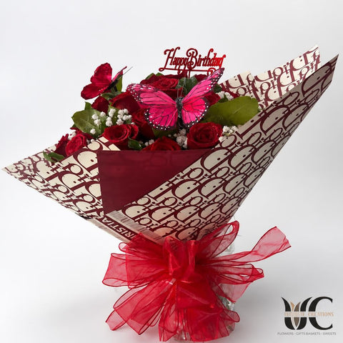 Wrapped bouquet of red roses with white filler flowers, greenery, pink butterfly decorations, a Happy Birthday topper, patterned paper, and a large sheer red bow.