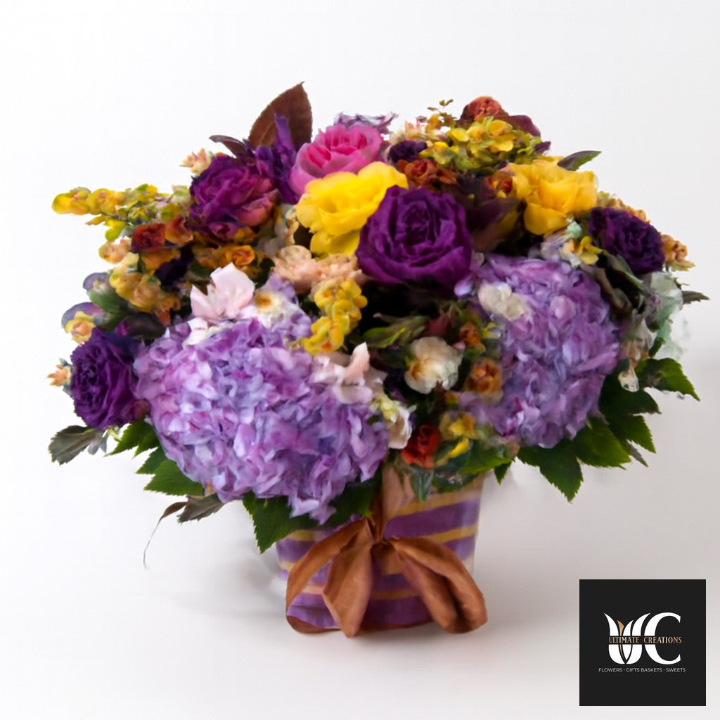 A colorful flower arrangement with purple, lavender, pink, and yellow blooms in a ribbon-tied decorative container.