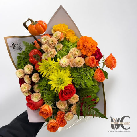 Lush hand-tied bouquet with orange seasonal accents, red roses, yellow flowers, cream blooms, green hydrangea, and fern foliage