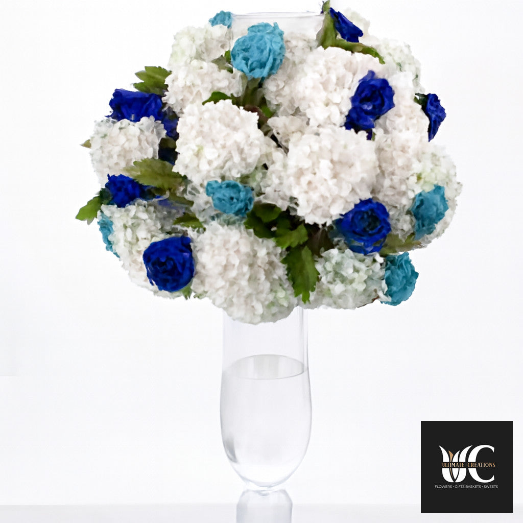 White hydrangea arrangement with blue accent flowers in a tall clear glass vase.