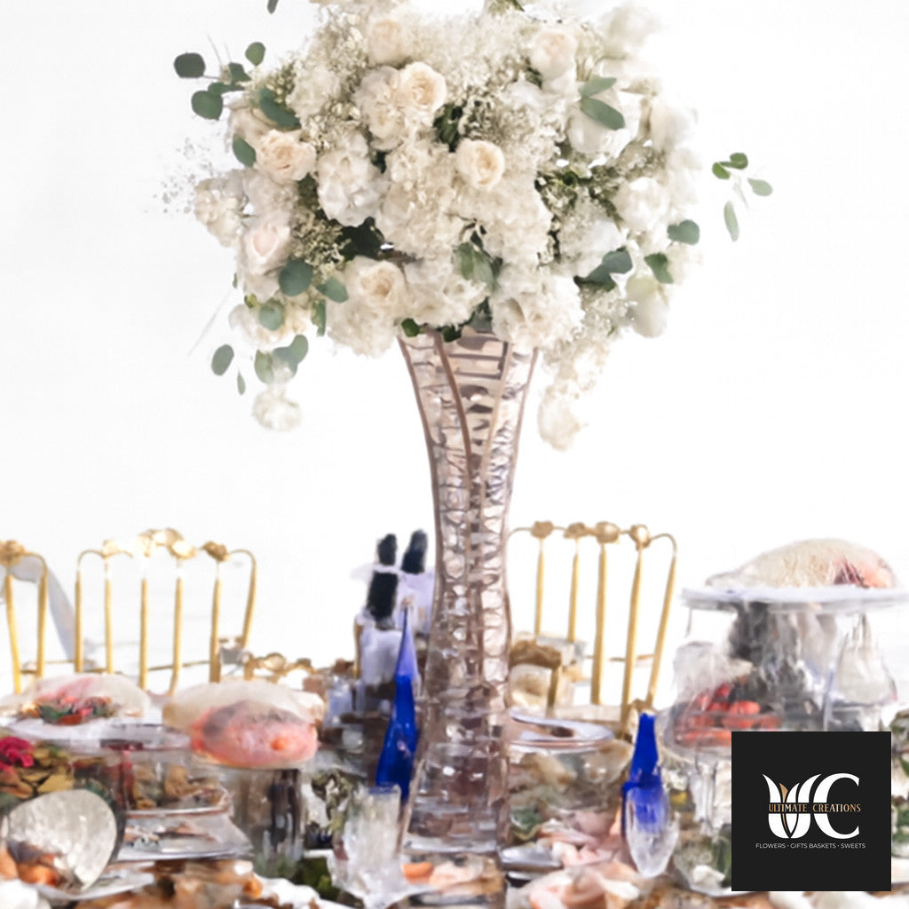 Tall crystal vase holding a large white floral arrangement with greenery on a decorated banquet table.
