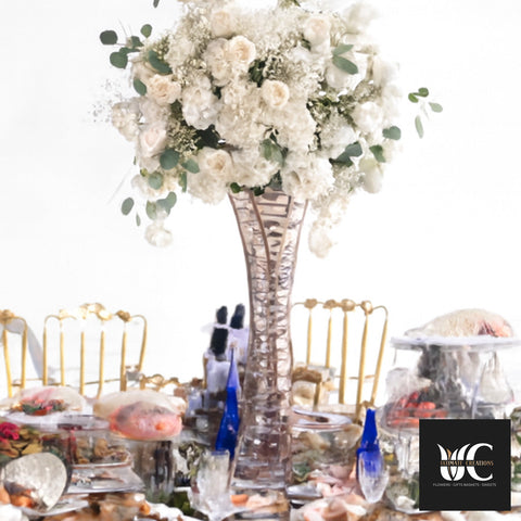 Tall crystal vase holding a large white floral arrangement with greenery on a decorated banquet table.