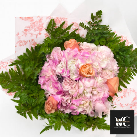 Hand-tied bouquet of pink, blush, peach, and white flowers with fern greenery wrapped in patterned paper.