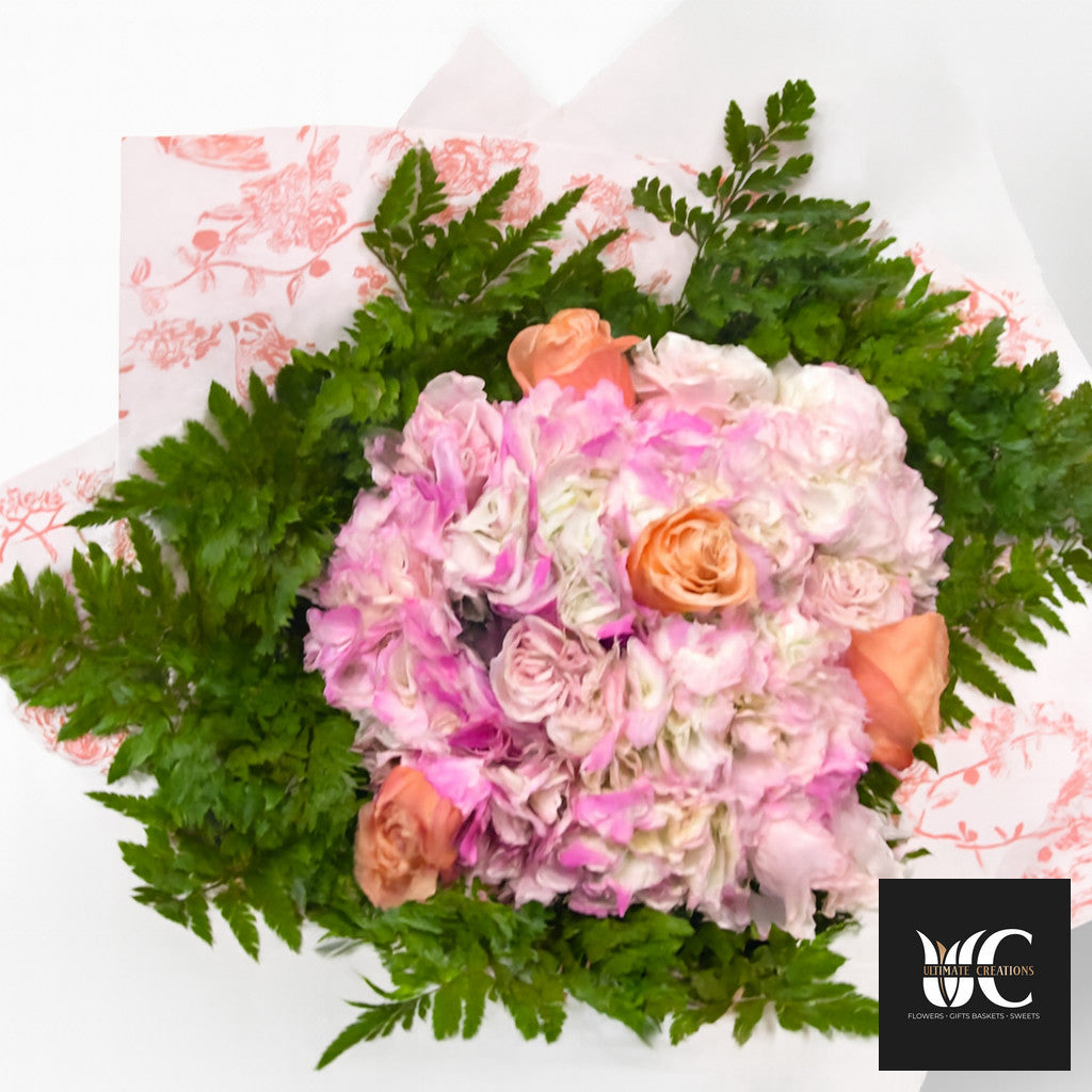 Hand-tied bouquet of pink, blush, peach, and white flowers with fern greenery wrapped in patterned paper.