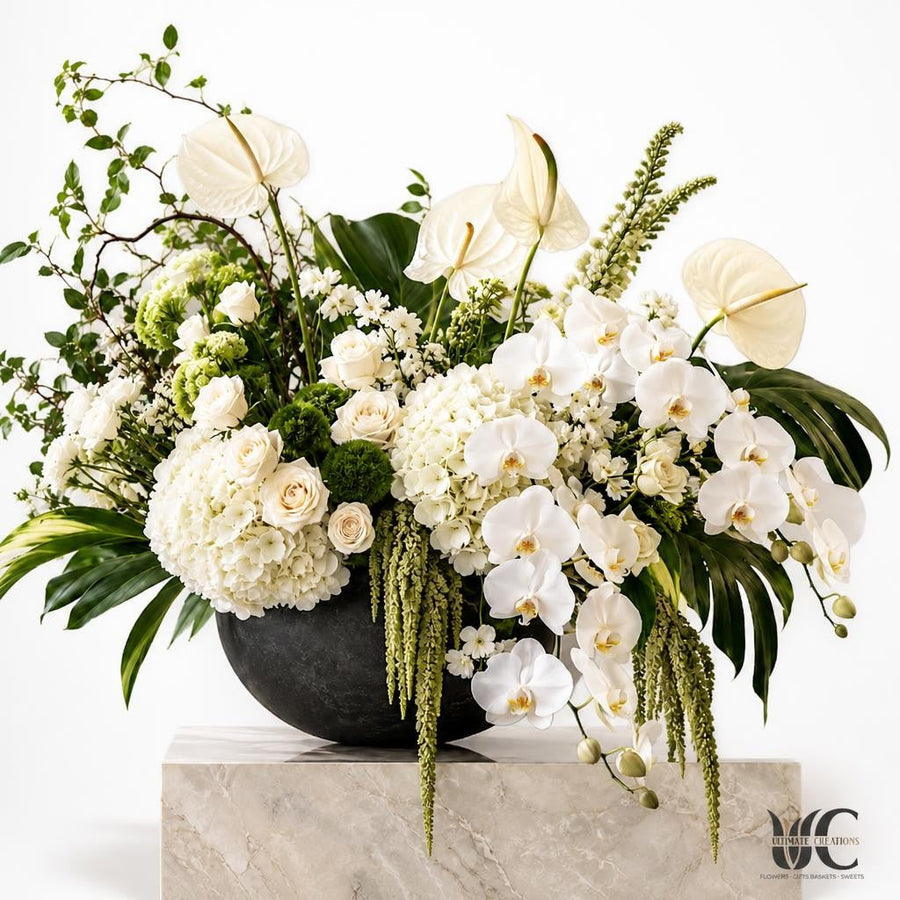 Modern white and green floral arrangement with orchid-like blooms, broad tropical leaves, rounded flowers, and trailing greenery in a charcoal bowl.