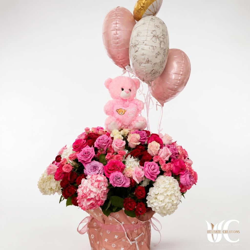 Pink and lavender flower arrangement in a wrapped container topped with a teddy bear and pink, white, and gold balloons.