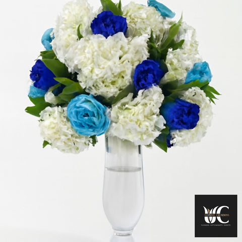 Tall glass vase holding a white and blue floral arrangement with greenery.