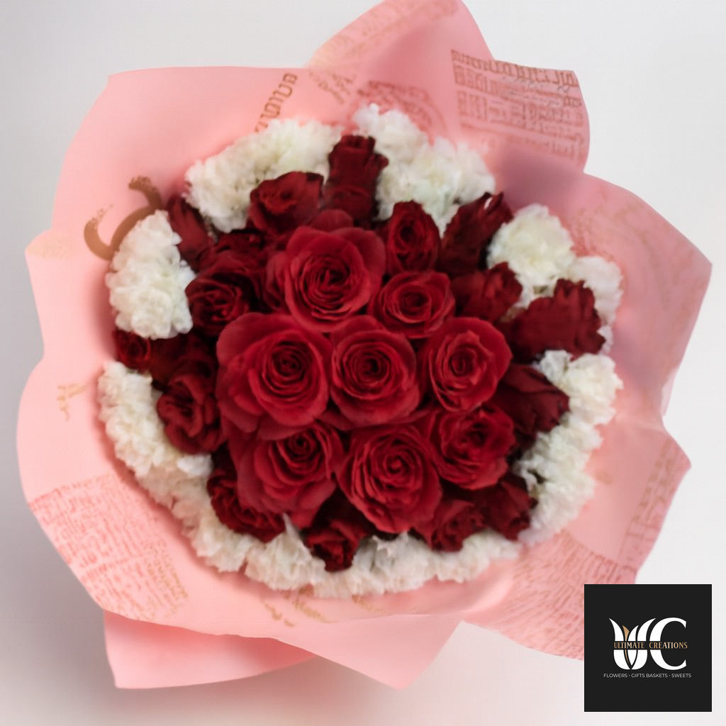 Round bouquet of red roses with white accent flowers wrapped in pink paper.