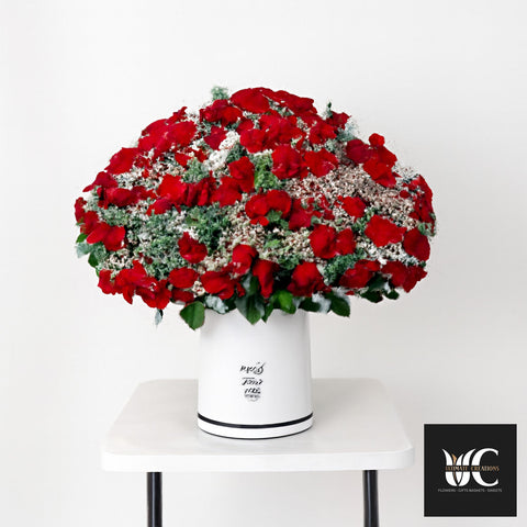 Large dome-shaped red flower arrangement with white filler and greenery in a white cylinder vase.