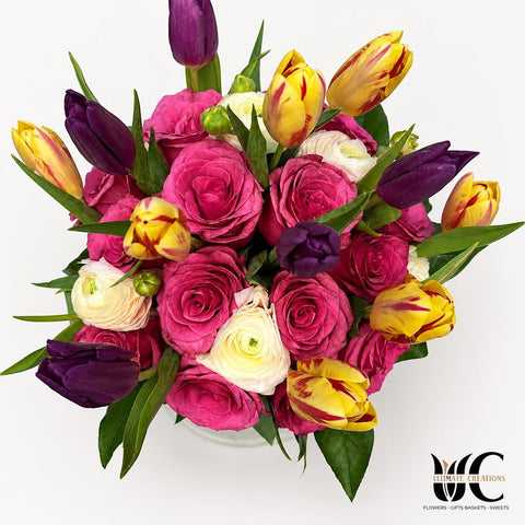 Full bouquet of pink roses, purple and yellow tulips, pale layered flowers, and green foliage arranged on a white surface.