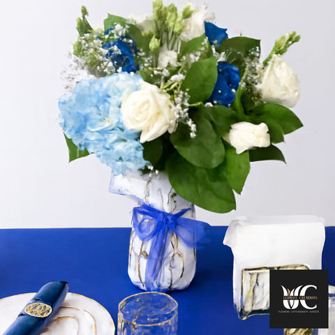 Blue and white floral centerpiece in a white marbled vase with a blue ribbon, featuring hydrangea, roses, and greenery.