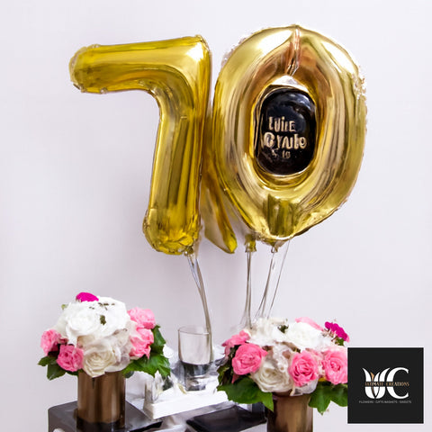 Gold number 70 balloons above pink and white floral arrangements in metallic vases.