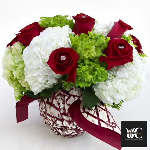Bouquet of red roses and white and green hydrangea arranged in a decorative vase with burgundy ribbon.