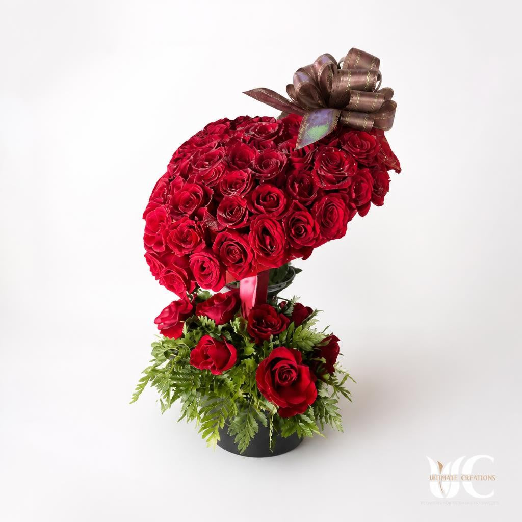 Dense red rose arrangement with a metallic bow, lower red roses, and green fern-like foliage on a store shelving unit.